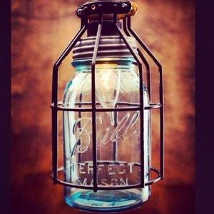 May include: A black metal cage light fixture with a clear glass mason jar. The jar has the text "Ball Perfect Mason" on it. The light is on and illuminating the jar.