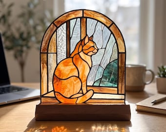 Op de afbeelding: Een boogvormig glas-in-loodpaneel met een oranje kat die in een raam zit. De kat is weergegeven in warme amberkleurige tinten, tegen een achtergrond van getextureerde blauwe en witte glaspanelen. Het stuk staat op een houten voet.