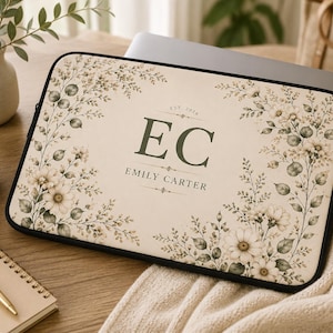 Puede incluir: Una funda rectangular para portátil con fondo color crema que presenta un estampado botánico de flores y hojas en tonos verde apagado y blanco. El centro muestra las iniciales EC en verde oscuro, el nombre EMILY CARTER y el texto EST. 2026.