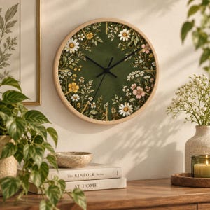 May include: A round wall clock with a light wood frame and a dark olive green face. The clock face features a botanical print of white daisies, yellow wildflowers, and pink blossoms arranged around the perimeter. It has three thin black hands.