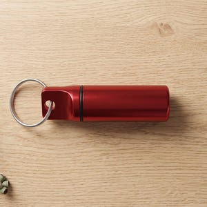 May include: A cylindrical red metal keyring capsule with a smooth finish and a black rubber O-ring seal. The capsule features a flat, extended end with a circular hole, through which a silver-toned metal split ring is securely attached.