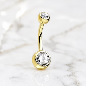 May include: A gold-toned curved barbell navel ring featuring two clear, round-cut faceted stones set in polished metal bezels. The larger stone is positioned at the base, while a smaller stone sits at the top of the curved metal post.