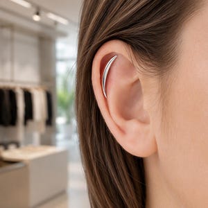 Puede incluir: Un ear cuff de tono plateado pulido con forma de media luna delgada y curva. La pieza metálica descansa a lo largo del cartílago superior de la oreja, presentando una superficie lisa y reflectante que se estrecha en puntas finas en ambos extremos.