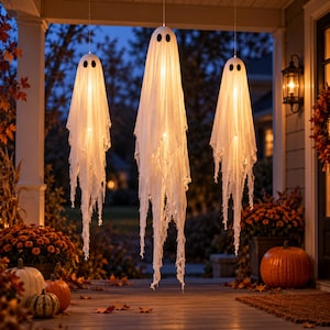 Puede incluir: Tres decoraciones de fantasmas colgantes hechas de tela blanca con bordes deshilachados y ojos ovalados negros. Cada fantasma está iluminado desde el interior por una luz cálida, creando un efecto brillante. Las figuras están suspendidas en el aire, con largas tiras de tela deshilachadas que cuelgan hacia el suelo.