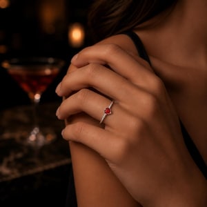 Puede incluir: Un anillo de plata con una pequeña piedra preciosa roja en forma de corazón montada sobre una banda de metal fina y pulida. El anillo se lleva en un dedo, destacando el color rojo vibrante de la piedra y el brillo metálico de la banda.
