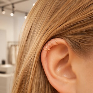 May include: A gold-toned ear cuff featuring a multi-band design that wraps around the upper cartilage of the ear. The piece consists of four thin, smooth wire bands, with the bottom two bands crossing over each other in an X-shaped pattern.