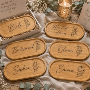 May include: A collection of six oval bamboo trays featuring laser-engraved floral illustrations and personalized text including Bride, Maid of Honor, Bridesmaid, Olivia, Sophia, and Emma, with one tray presented in an open white gift box.