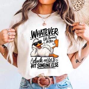 May include: A cream-colored t-shirt featuring a graphic of a white duck wearing black sunglasses, reclining with one orange foot raised. The text reads: WHATEVER LIFE THROWS AT YOU, duck and Let It HIT SOMEONE ELSE in black stylized lettering.