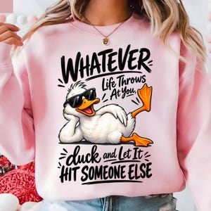 May include: A light pink sweatshirt featuring a graphic of a white duck wearing black sunglasses, reclining with one leg raised. The text reads: WHATEVER Life Throws AT YOU, duck and Let It HIT SOMEONE ELSE in black, stylized, handwritten-style font.