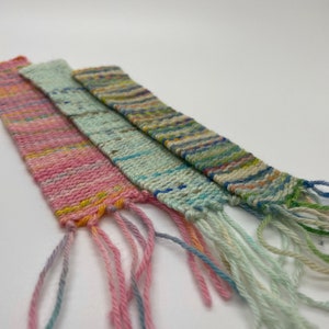 May include: Four colorful woven bookmarks with fringe. The bookmarks are in shades of pink, blue, and green, with flecks of yellow and other colors. The fringe is made of matching yarn.