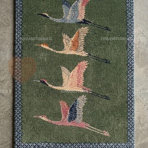 May include: A rectangular green rug featuring a repeating pattern of five flying cranes in shades of pink, orange, and blue. The rug has a dark blue patterned border and a white fringe along the bottom edge. The text ZIVAANSTUDIOS is visible.