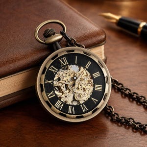 May include: A bronze-toned mechanical pocket watch featuring a black dial with gold-colored Roman numerals and an exposed skeleton movement. The watch includes a matching metal chain attached to the top crown and is designed with a circular case.