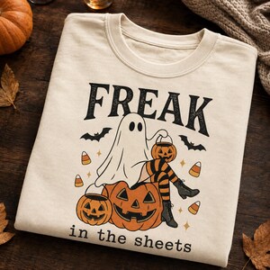 Puede incluir: Una camiseta de cuello redondo color crema con un gráfico de un fantasma sentado sobre una calabaza grande. El fantasma lleva medias a rayas naranjas y negras y botas, sosteniendo un pequeño cubo de calabaza. El texto dice FREAK in the sheets.