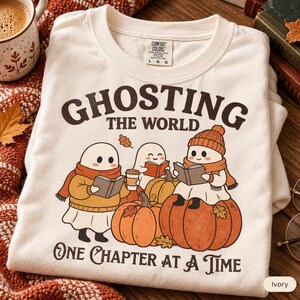 May include: An ivory crewneck sweatshirt featuring a graphic of three ghosts reading books and holding coffee among pumpkins. The text reads GHOSTING THE WORLD ONE CHAPTER AT A TIME. The garment has a Comfort Colors brand label at the inner neckline.