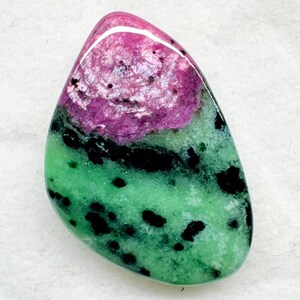 Può includere: Una gemma di zoisite lucidata a forma di goccia con un'inclusione di rubino magenta brillante nella parte superiore, contrastata da una base verde brillante punteggiata da inclusioni minerali di orneblenda nera sulla superficie liscia e lucida.
