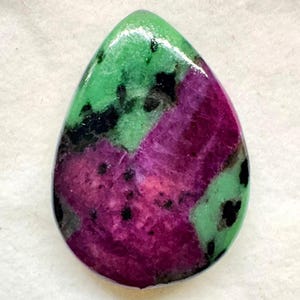 Peut inclure: Un cabochon de pierre précieuse poli en forme de poire présentant un mélange vibrant de couleurs magenta profond et vert lime vif, accentué par des inclusions minérales noires dispersées sur toute la surface lisse, brillante et arrondie.