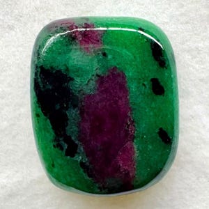 Può includere: Una pietra preziosa lucida di forma rettangolare con angoli arrotondati, caratterizzata da una base di colore verde vibrante con macchie irregolari e prominenti di magenta intenso e inclusioni nere scure distribuite sulla superficie liscia e riflettente.