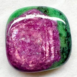 Puede incluir: Una piedra preciosa pulida y redondeada que presenta un centro rosa magenta vibrante rodeado por un borde verde bosque profundo. La superficie es lisa y reflectante, mostrando texturas cristalinas naturales en las secciones minerales.