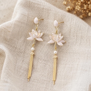 May include: A pair of gold-tone drop earrings featuring a white lotus flower design with a small white bead accent and a dangling gold-tone chain tassel. The earrings have a post-back closure and a delicate, elongated silhouette with metallic finish.