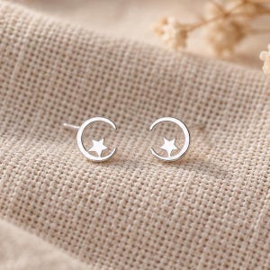 May include: A pair of silver-toned stud earrings, each featuring a crescent moon shape with a small five-pointed star centered within the curve. The earrings have a polished metallic finish and are designed as simple, symmetrical celestial motifs.