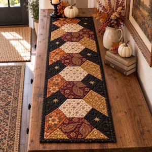 May include: A rectangular quilted table runner featuring a repeating hexagonal pattern. The design incorporates various fabric prints in shades of black, burnt orange, mustard yellow, and cream, including floral motifs, paisley patterns, and small star accents.