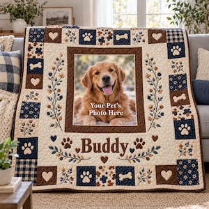 May include: A cream-colored quilt featuring a central photo frame with the text Your Pet's Photo Here, surrounded by a border of navy blue and brown squares with paw prints, bones, and hearts. The name Buddy is printed below the frame.