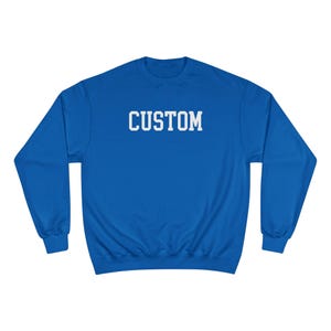 May include: A royal blue long-sleeve crewneck sweatshirt featuring the word CUSTOM printed in white block capital letters across the center of the chest. The garment has a ribbed collar, cuffs, and hem, with a standard pullover design.
