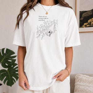 Op de afbeelding: Een wit t-shirt met korte mouwen en een zwarte lijntekening van een auto op een weg omzoomd door palmbomen. De tekst boven de illustratie luidt, There are no wrong roads to anywhere. Het t-shirt heeft een ronde hals en een losse pasvorm.