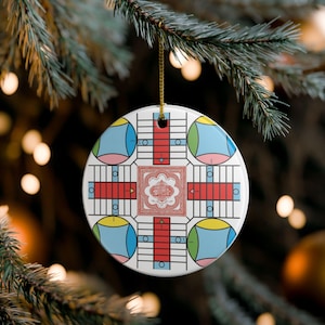 May include: A round white ceramic ornament featuring a colorful board game design. The graphic includes four quadrants with red, yellow, blue, and green accents, black grid lines, and a central square with intricate red and white decorative patterns.