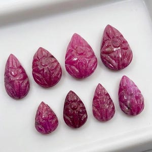Peut inclure: Huit pierres précieuses en forme de goutte d'une teinte magenta-rose profond, chacune présentant un motif floral sculpté en surface. Les pierres sont disposées en deux rangées sur un plateau blanc, montrant des tailles variées.