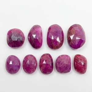Puede incluir: Una colección de nueve rubíes facetados de color rojo rosado intenso dispuestos en dos filas. Cada piedra preciosa presenta una forma ovalada o redondeada irregular y pulida con facetas geométricas visibles que reflejan la luz.