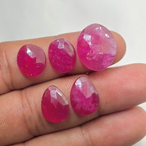 Puede incluir: Cinco piedras preciosas facetadas de un vibrante color rojo rosado en varias formas. Tres piedras son de forma ovalada, mientras que dos tienen forma de pera. Cada piedra presenta una superficie pulida y multifacetada que refleja la luz.