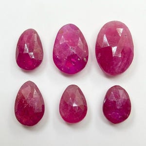 Puede incluir: Seis piedras preciosas de rubí facetadas en forma de pera dispuestas en dos filas. Cada piedra muestra un tono rojo magenta intenso con una superficie pulida de múltiples facetas que refleja la luz, mostrando variaciones naturales de color.