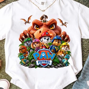 Puede incluir: Una camiseta blanca con un estampado gráfico colorido de los personajes de Paw Patrol y un dinosaurio grande. El diseño incluye el logotipo azul y amarillo de Paw Patrol, varios cachorros de dibujos animados con uniforme y criaturas prehistóricas.