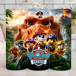 Op de afbeelding: Een cilindervormige beker met een levendige Paw Patrol-afbeelding met diverse tekenfilmpuppy's in een prehistorische jungleomgeving. Het ontwerp bevat een grote dinosaurus, een vulkaan, watervallen en het blauw-rode Paw Patrol-logo.