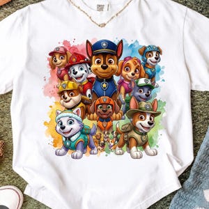 Può includere: Una t-shirt bianca a maniche corte con una grafica colorata in stile acquerello che raffigura l'insieme dei personaggi di Paw Patrol. Il design mostra diversi cuccioli animati nelle loro uniformi e attrezzature caratteristiche, disposti in gruppo su uno sfondo di vivaci schizzi di vernice multicolore.