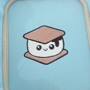 Op de afbeelding: Een borduurontwerp op lichtblauwe stof met een s'more-figuurtje met een wit marshmallow-midden, bruine graham cracker-boven- en onderkant en een kleine donkerbruine chocoladedruppel, vastgezet in een rechthoekige borduurring.