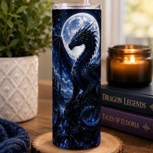 May include: A cylindrical tumbler with a clear lid and straw, featuring a dark blue graphic print of a dragon perched on a rocky cliff against a large, bright full moon, surrounded by a starry night sky and glowing blue crystal formations.
