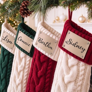 May include: Four cable-knit Christmas stockings hanging in a row. The stockings are colored dark green, white, dark red, and white. Each features a rectangular beige name tag with the names Lisa, Connie, Bertha, and Sidney in black script.