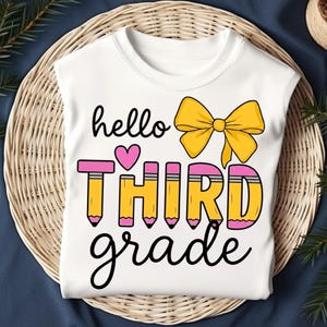 Peut inclure: Un t-shirt blanc avec un motif graphique comprenant le texte "hello" en noir, un nœud jaune, un cœur rose, le mot "THIRD" formé par des crayons jaunes à gommes roses, et le mot "grade" écrit en lettres cursives noires.