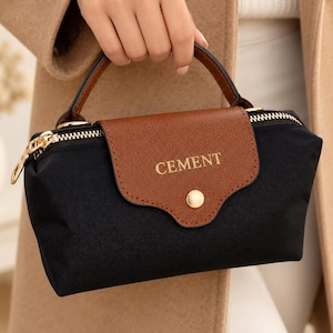 Puede incluir: Un bolso de mano pequeño de color negro con una solapa marrón que presenta la palabra CEMENT en letras doradas. El bolso tiene un cierre de broche de metal dorado, un asa superior y una cremallera dorada en el borde superior.