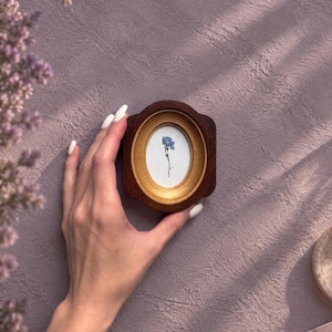 May include: A small, oval-shaped picture frame featuring a dark brown wooden outer border and a gold-toned inner rim. The frame contains a white background with a single, delicate blue flower illustration centred within the oval display area.