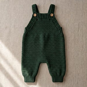 May include: A dark forest green crochet overall featuring a ribbed neckline and shoulder straps secured with two small light brown wooden buttons. The garment is crafted with a consistent horizontal stitch pattern and ribbed cuffs at the ankles.