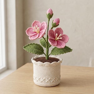 以下が含まれることがあります： 白い雄しべを持つ2つの開いたピンクの花と、緑の茎についた2つの閉じたピンクの蕾が特徴の、かぎ針編みの鉢植えです。質感のある緑の葉を持ち、縁がスカラップ状で底にクロスステッチ模様が施されたクリーム色のかぎ針編みの鉢に入っています。