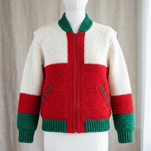 May include: A long-sleeve crochet bomber jacket featuring a color-blocked design in cream, red, and green. The jacket has a ribbed collar, cuffs, and hem, a central metal zipper closure, and two front pockets with green trim on the red panels.