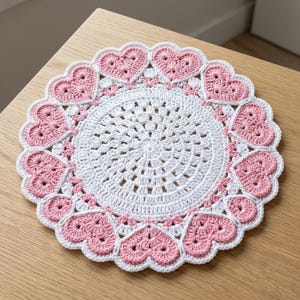 May include: A round, white crocheted doily featuring a central circular pattern with an intricate, openwork design. The outer edge is decorated with a series of repeating, soft pink crocheted heart motifs arranged in a scalloped, circular border.