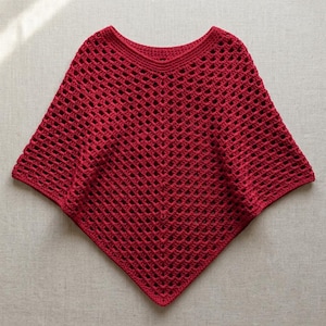 Peut inclure: Un poncho en crochet rouge profond présentant un motif ajouré en forme de losange sur tout le corps. Le vêtement possède une large encolure arrondie avec une bordure cousue solide et un ourlet pointu créant une silhouette triangulaire.