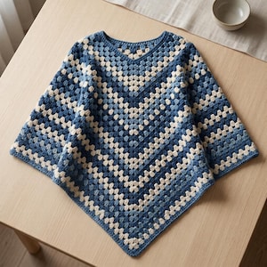Op de afbeelding: Een gehaakte poncho met een V-hals en een geometrisch granny square patroon. Het kledingstuk is gemaakt met afwisselende rijen van crème, lichtblauw en donker marineblauw garen, wat een symmetrisch chevronontwerp vormt dat naar beneden wijst.