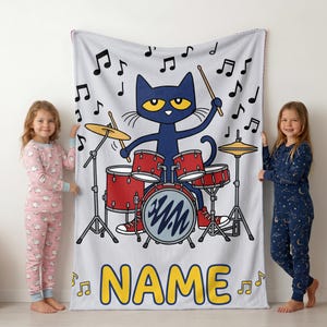 May include: A light gray fleece blanket featuring a cartoon blue cat playing a red drum set with yellow cymbals. Black musical notes float around the cat. The word NAME is printed in large yellow letters at the bottom of the blanket.