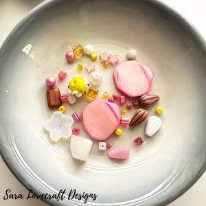 May include: A collection of colorful beads and decorative elements in a ceramic bowl. The assortment includes pink, yellow, and clear beads, along with flower and star-shaped accents. The text "Sara Lovecraft Designs" is at the bottom.